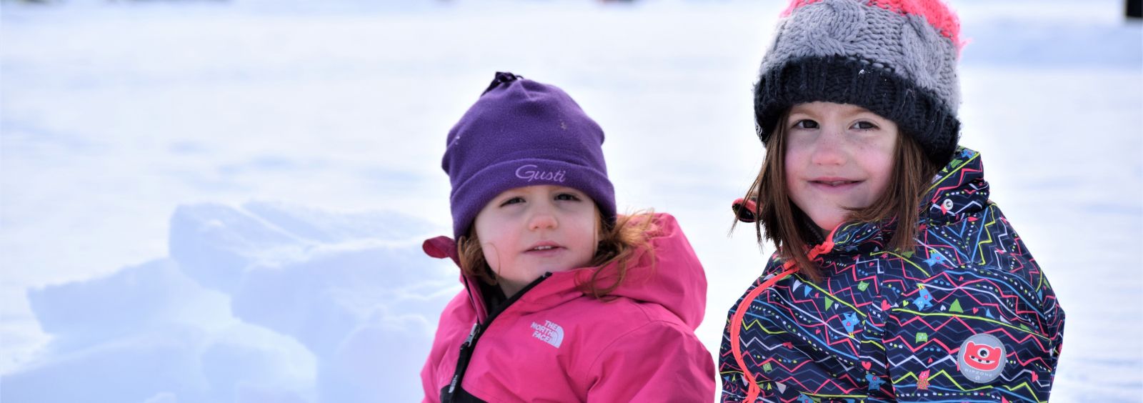 Two young children smiling in winter clothing with snow behind them.