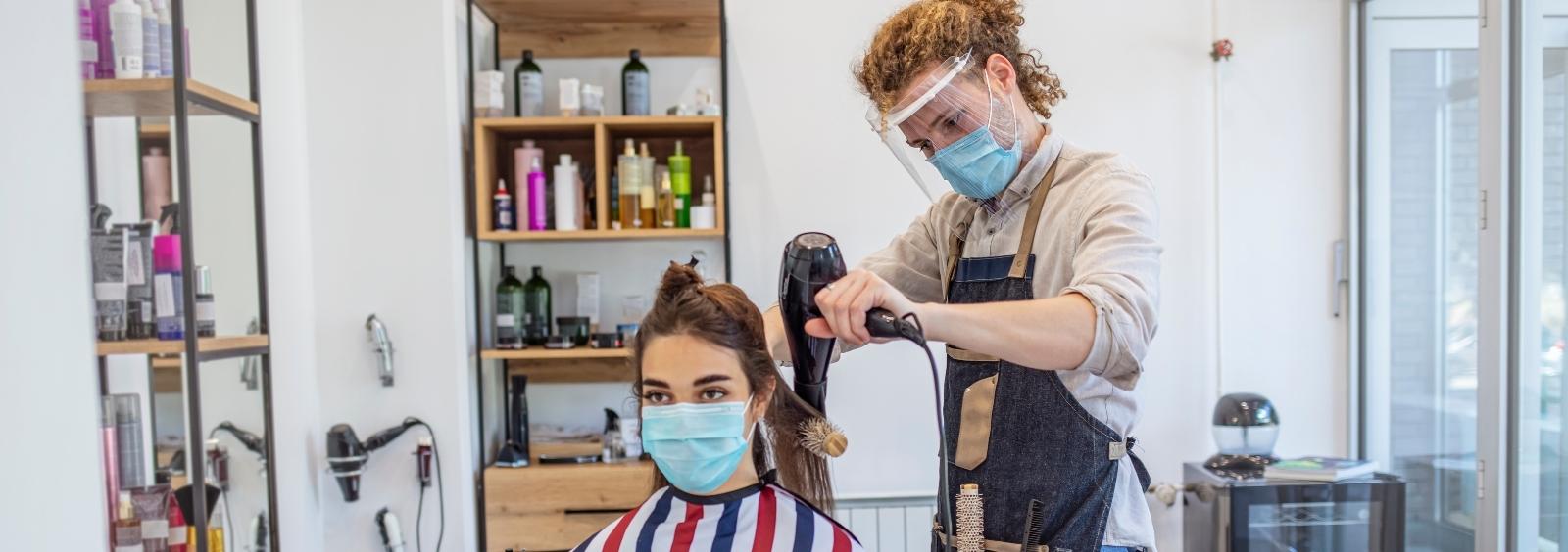 A barber cuts hair on a women with masks and a face shield on. 