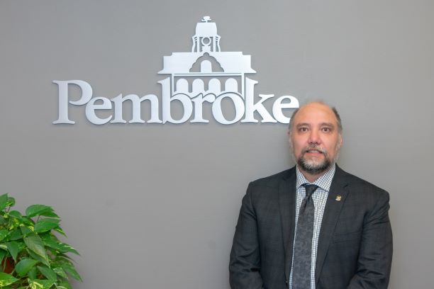 Pembroke mayor Ron Gervais standing in front of the logo of the City of Pembroke on a grey wall.