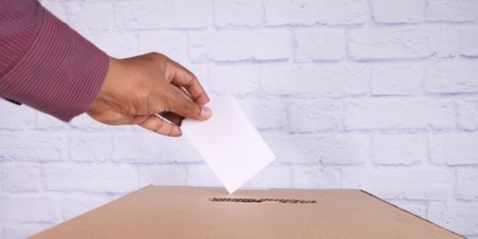 A hand places a ballot in a ballot box.