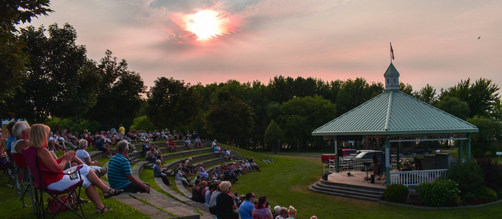 Wide shot of the John Priebe Band playing at the Riverwalk Amphitheatre. Crowd fills the Amphitheatre seating to the right.